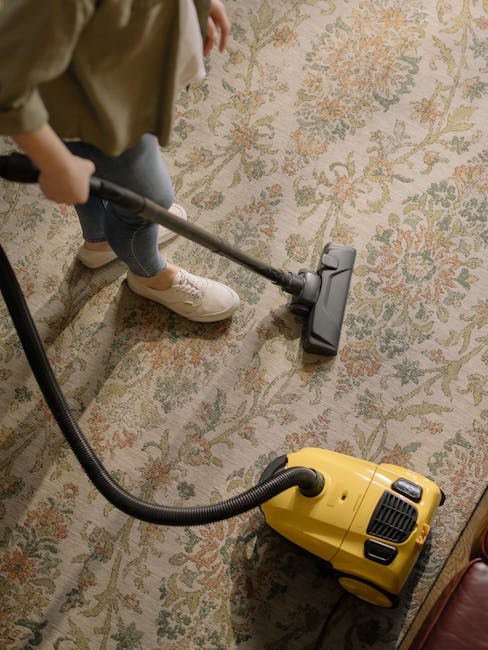 Close-up of a vacuum cleaner attachment in use on a patterned area rug with floral and leaf motifs in beige, green, and orange tones. The vacuum head is black with a small switch visible, and it is po